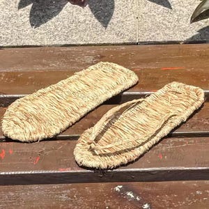 May include: A pair of traditional Japanese straw sandals, or waraji, made from woven straw. The sandals are light brown in color and feature a simple thong design. They are placed on a wooden surface.