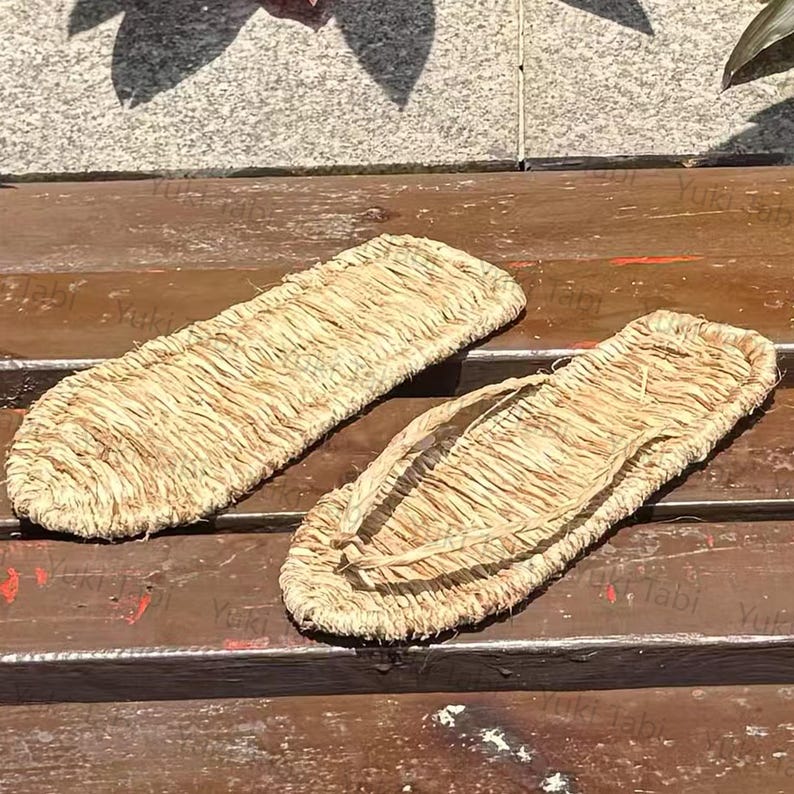 May include: A pair of traditional Japanese straw sandals, or waraji, made from woven straw. The sandals are light brown in color and feature a simple thong design. They are placed on a wooden surface.