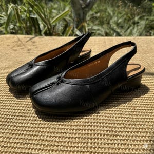 May include: A pair of black Tabi shoes with a split toe design. These leather slingback sandals have a low heel and a small bow detail on the vamp. The shoes are displayed on a woven surface.