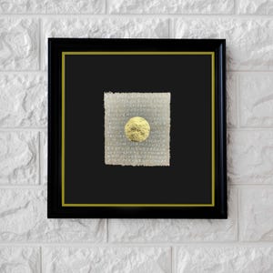 May include: A framed artwork featuring a textured, off-white square with a gold-colored sphere at its center. The artwork is set against a black background within a black frame with a gold inner border. The background is a white brick wall.