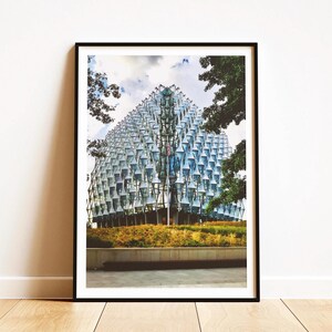 Puede incluir: Impresión enmarcada de un edificio moderno con una fachada geométrica única. El exterior del edificio presenta numerosos paneles de vidrio, que reflejan el cielo. La imagen está enmarcada en negro, con una vista de árboles y una zona de césped en primer plano.