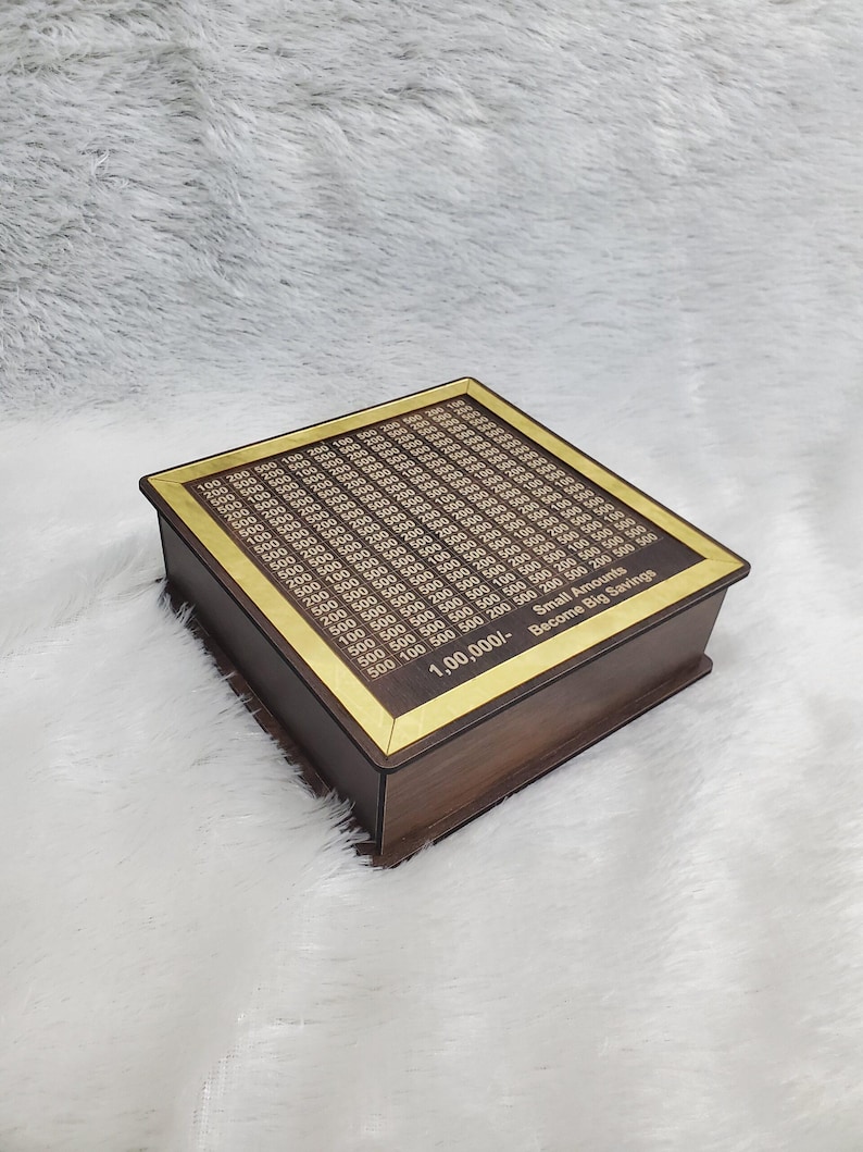 Puede incluir: Una caja de ahorros de madera con un borde dorado. La parte superior est&aacute; grabada con un patr&oacute;n de cuadr&iacute;cula y n&uacute;meros. El texto "Small Amounts Become Big Savings" es visible en el lateral.