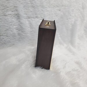 Puede incluir: Una caja de madera marr&oacute;n, dise&ntilde;ada para parecerse a un libro, con una cerradura dorada en la parte superior. La caja es rectangular y tiene un acabado marr&oacute;n oscuro. El fondo es una superficie blanca y esponjosa.