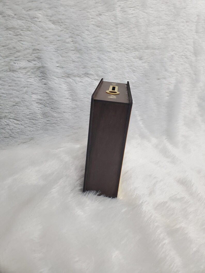 Puede incluir: Una caja de madera marr&oacute;n, dise&ntilde;ada para parecerse a un libro, con una cerradura dorada en la parte superior. La caja es rectangular y tiene un acabado marr&oacute;n oscuro. El fondo es una superficie blanca y esponjosa.