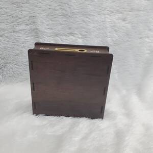 Puede incluir: Una caja de madera marr&oacute;n oscuro con una ranura en la parte superior y una peque&ntilde;a abertura circular. La caja tiene forma rectangular y est&aacute; sobre una superficie blanca y esponjosa. La caja parece ser un contenedor de almacenamiento o un art&iacute;culo decorativo.