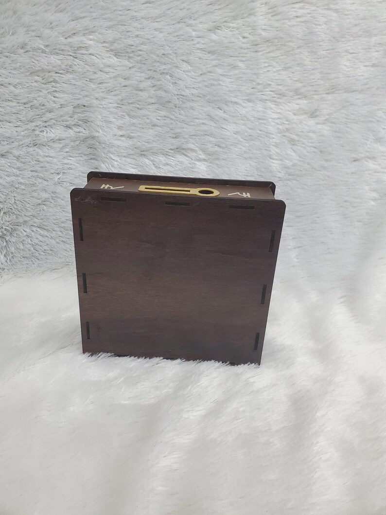 Puede incluir: Una caja de madera marr&oacute;n oscuro con una ranura en la parte superior y una peque&ntilde;a abertura circular. La caja tiene forma rectangular y est&aacute; sobre una superficie blanca y esponjosa. La caja parece ser un contenedor de almacenamiento o un art&iacute;culo decorativo.