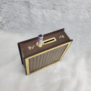 Puede incluir: Un banco de dinero de madera con forma de libro, con una ranura para insertar billetes. El banco tiene un exterior marr&oacute;n oscuro con adornos dorados y una parte delantera estampada. Se ve un billete enrollado en la ranura.