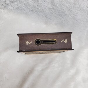 Puede incluir: Una caja rectangular de madera con exterior marr&oacute;n oscuro. La caja tiene una ranura y un agujero circular en la parte delantera. Hay dos s&iacute;mbolos en la parte delantera de la caja. La caja est&aacute; sobre una superficie blanca y esponjosa.