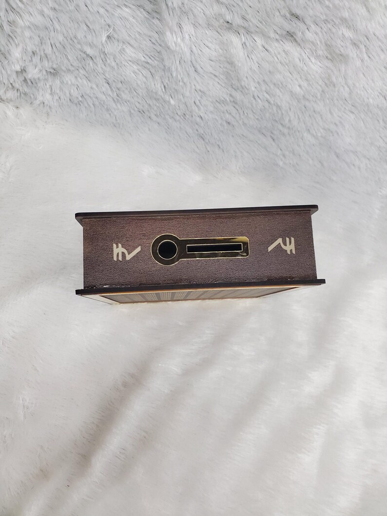 Puede incluir: Una caja rectangular de madera con exterior marr&oacute;n oscuro. La caja tiene una ranura y un agujero circular en la parte delantera. Hay dos s&iacute;mbolos en la parte delantera de la caja. La caja est&aacute; sobre una superficie blanca y esponjosa.