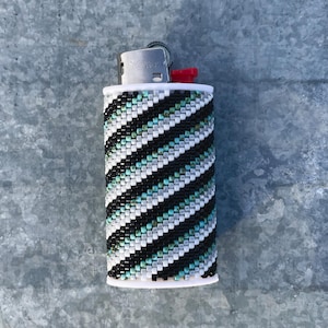 Pode incluir: Um isqueiro com uma capa de missangas com um padrão de riscas diagonais. As missangas estão dispostas em filas alternadas de preto, branco, cinzento e azul-petróleo. O topo do mecanismo metálico do isqueiro é visível com um botão vermelho.