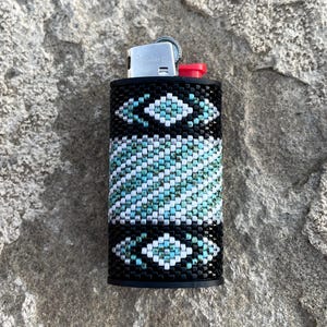 May include: A cylindrical lighter case featuring a beaded exterior with a geometric pattern in black, white, and turquoise. The design includes diamond shapes at the top and bottom with a central band of diagonal stripes. A silver metal lighter is inserted.