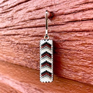 Könnte beinhalten: Ein rechteckiger Ohrring mit einem silbernen Haken. Der Ohrring besteht aus kleinen, farbigen Perlen in einem Muster aus Weiß, Schwarz und Rot. Der Ohrring hängt vor einer verwitterten, rotbraunen Holzoberfläche.