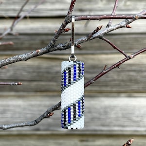 Könnte beinhalten: Ein zylindrischer Perlenanhänger mit silberfarbenem Verschluss hängt an einem Ast. Der Anhänger hat ein Spiraldesign aus weißen, schwarzen und blauen Perlen. Der Hintergrund ist eine verwitterte Holzoberfläche.