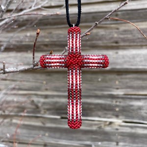 Puede incluir: Un colgante de cruz de cuentas con cuentas rojas, blancas y negras. La cruz está suspendida de un cordón negro, sobre un fondo de madera desgastada. La cruz es un símbolo religioso.