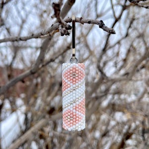 May include: A cylindrical beaded charm with a silver-coloured clasp. The charm features a spiral pattern of pink, white, and silver beads. The background is a blurred view of tree branches against a cloudy sky.