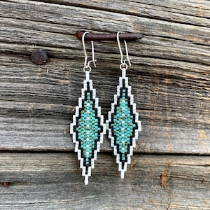 May include: A pair of long, diamond-shaped beaded earrings featuring a geometric pattern. The design is composed of small seed beads in white, black, and various shades of turquoise and teal. Each earring hangs from a silver-toned metal hook.