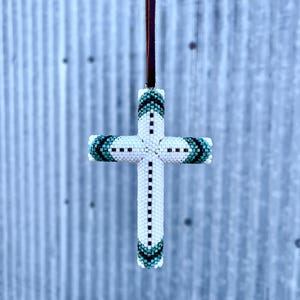 Può includere: Un ciondolo a forma di croce di perline con un cordino in pelle marrone. La croce è bianca con accenti turchesi e neri. La croce è fatta di piccole perline e ha un design dettagliato.