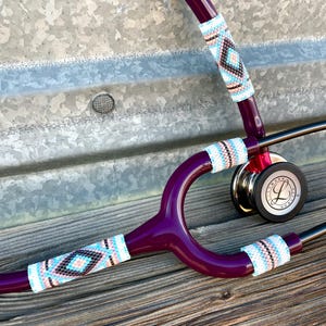 May include: A deep plum-coloured stethoscope featuring three sections of intricate, multi-coloured beaded covers. The beads display a geometric pattern in shades of turquoise, white, black, and soft pink. The chest piece is black with the Littmann logo.