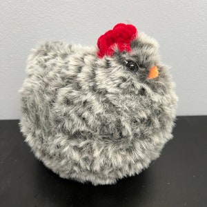 May include: A fluffy, round chicken toy with grey and white fur. It features a bright red comb and a small orange beak. The toy is designed to resemble a hen.