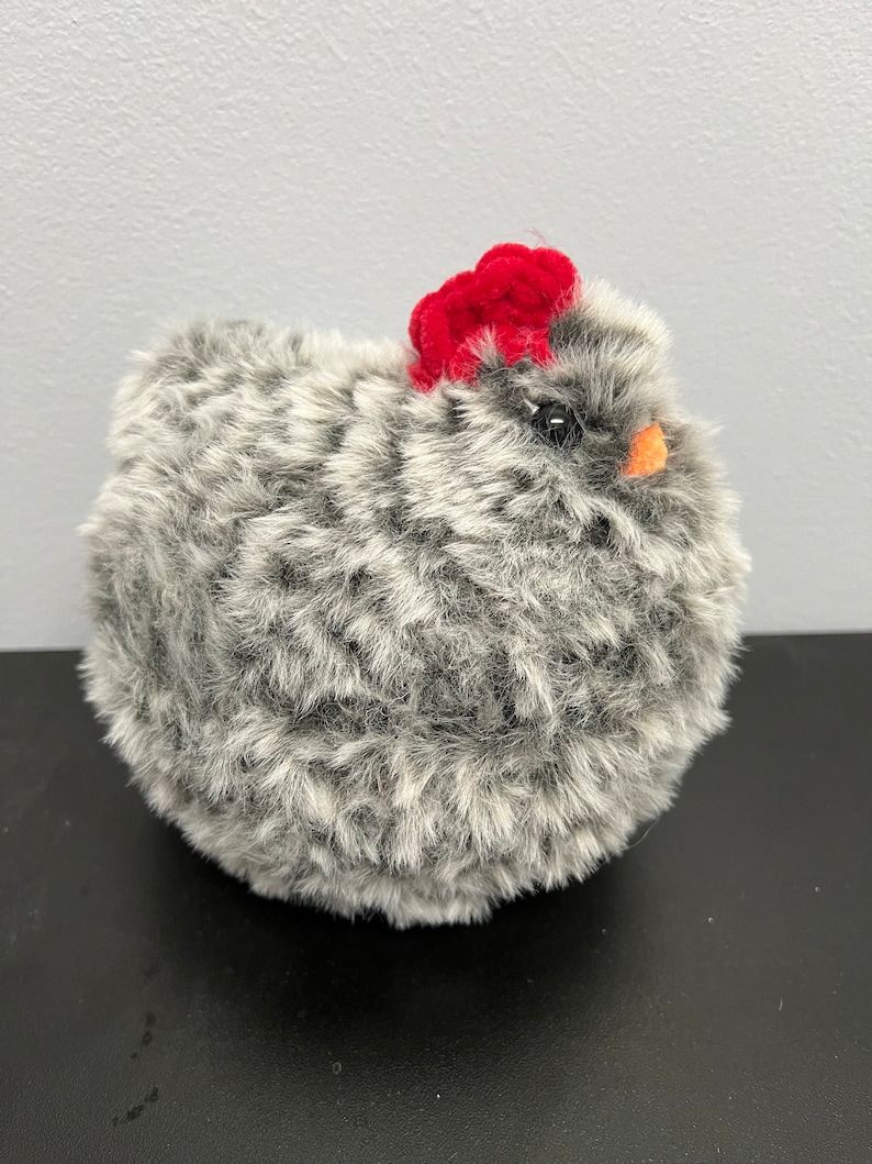 May include: A fluffy, round chicken toy with grey and white fur. It features a bright red comb and a small orange beak. The toy is designed to resemble a hen.