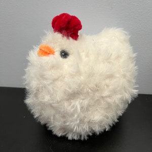 May include: A soft, round, white plush chicken toy with a vibrant red comb and an orange beak. The toy has a black eye. The chicken is a cuddly toy, sitting on a black surface.