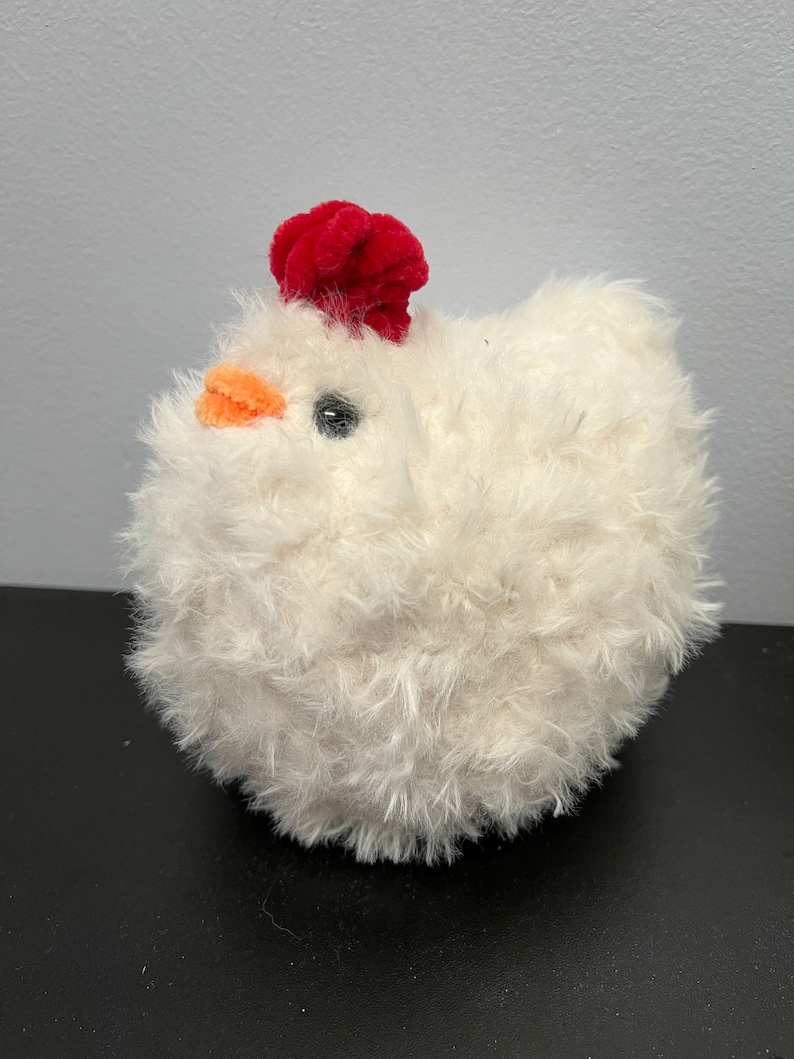 May include: A soft, round, white plush chicken toy with a vibrant red comb and an orange beak. The toy has a black eye. The chicken is a cuddly toy, sitting on a black surface.
