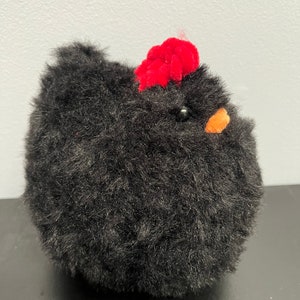 May include: A fluffy black chicken toy with a red comb and an orange beak. The soft toy has a round body and a textured appearance, with a small black bead for an eye. A charming decorative item.