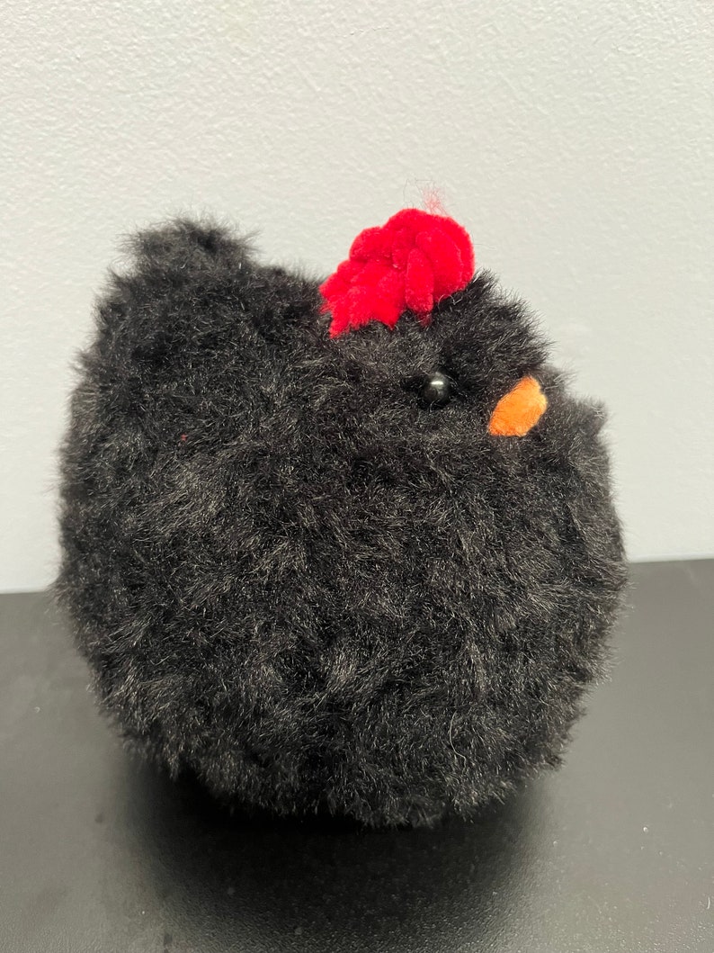 May include: A fluffy black chicken toy with a red comb and an orange beak. The soft toy has a round body and a textured appearance, with a small black bead for an eye. A charming decorative item.