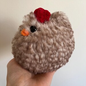 May include: A fluffy, round, brown chicken toy with a red heart-shaped comb and an orange beak. The chicken has a black eye and is made of soft material. The toy is held in a person's hand.