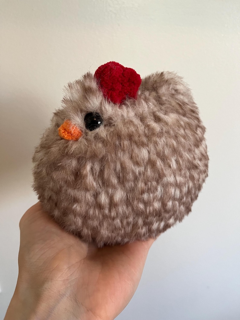 May include: A fluffy, round, brown chicken toy with a red heart-shaped comb and an orange beak. The chicken has a black eye and is made of soft material. The toy is held in a person's hand.