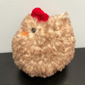 May include: A fluffy chicken toy with brown fur, a red comb, an orange beak, and a black eye. The stuffed animal is round and fluffy, resembling a chick. This soft toy is suitable for children.
