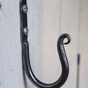 May include: A hand-forged, black metal hook mounted on a white wooden surface. The hook is attached with two silver screws. The hook's design features a curved body and a decorative, curled end, suitable for hanging items.