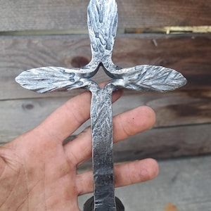 May include: Hand-forged metal cross with a textured, weathered appearance. The cross features a vertical stem and horizontal arms with rounded ends. The metal has a dark, gray color, and the cross is held in a hand.