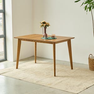 May include: A rectangular wooden dining table with four tapered legs and a smooth, light brown finish. A dark vase containing a bouquet of flowers and two small glass tumblers sit on a circular green placemat in the center of the table surface.
