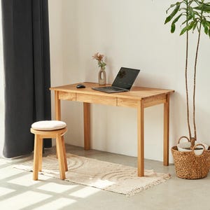 Könnte beinhalten: Ein heller Schreibtisch aus Holz mit zwei Schubladen, ein passender Holzhocker mit rundem weißem Kissen, ein schwarzer Laptop, eine schwarze Computermaus und eine kleine Keramikvase mit Trockenblumen auf der rechteckigen Tischoberfläche.