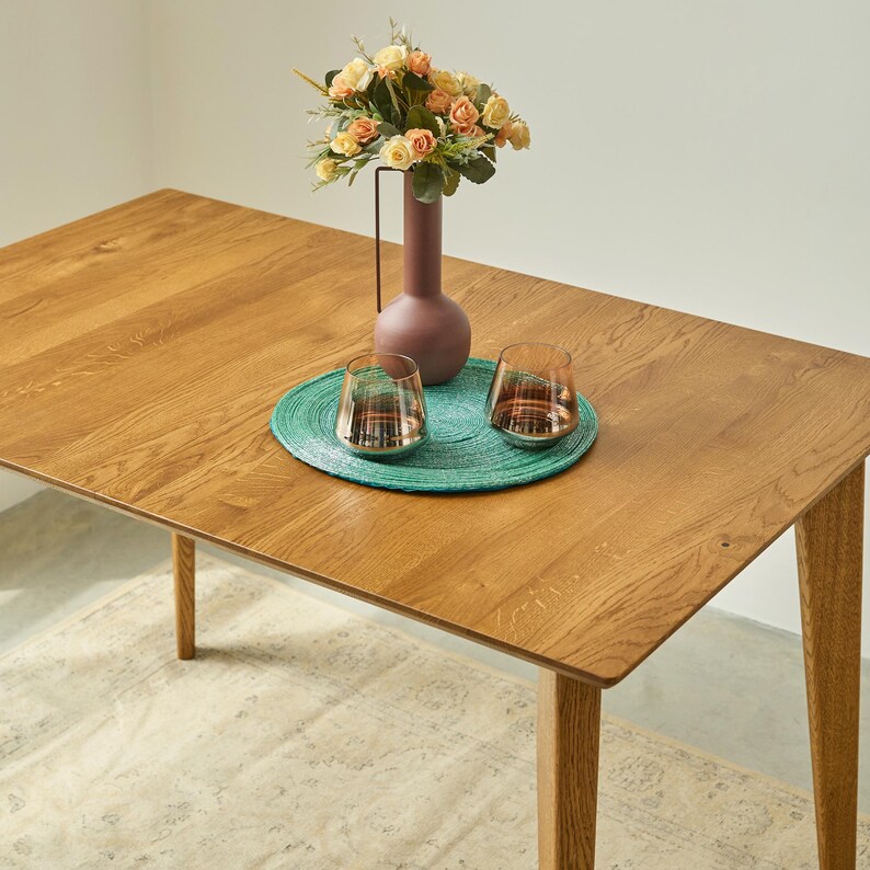 May include: A rectangular wooden dining table with a smooth grain finish. Centered on the table is a round teal woven placemat holding two clear glass tumblers and a tall, matte mauve vase filled with a bouquet of peach and cream artificial roses.