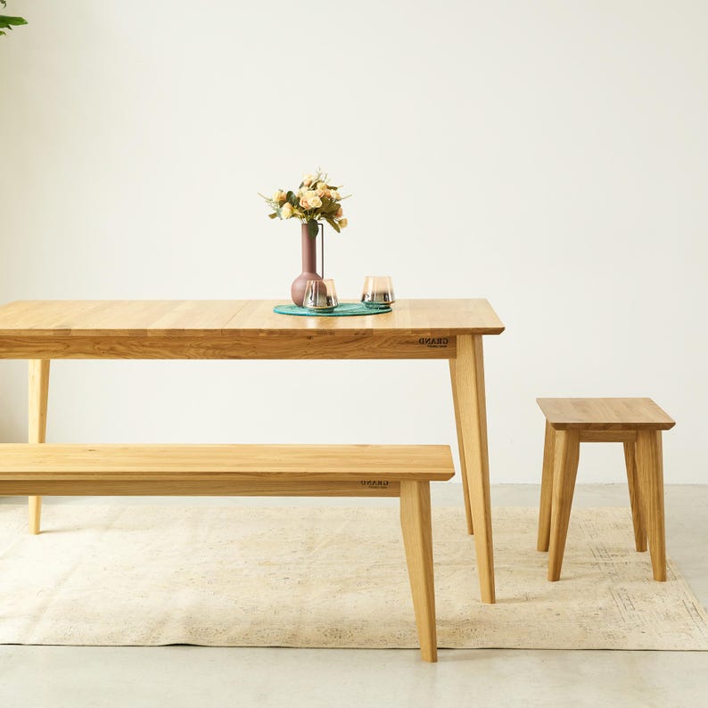 May include: A light-toned wooden dining set consisting of a rectangular table, a long bench, and a small stool. The table features the word GRAND printed on the side. A tall vase with flowers and two small glass containers sit on the table surface.