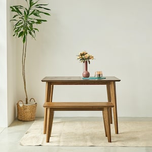 May include: A rectangular wooden dining table with four tapered legs and a matching wooden bench. On the table sits a tall, slender, mauve-colored vase containing a bouquet of peach and cream-colored flowers, alongside a small glass candle holder.