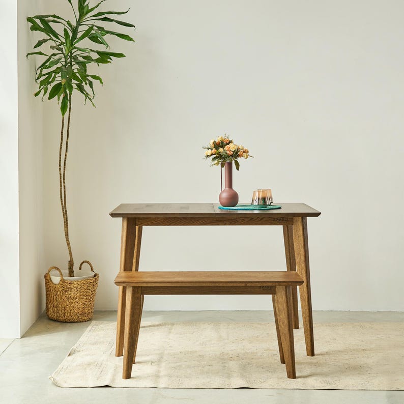 May include: A rectangular wooden dining table with four tapered legs and a matching wooden bench. On the table sits a tall, slender, mauve-colored vase containing a bouquet of peach and cream-colored flowers, alongside a small glass candle holder.