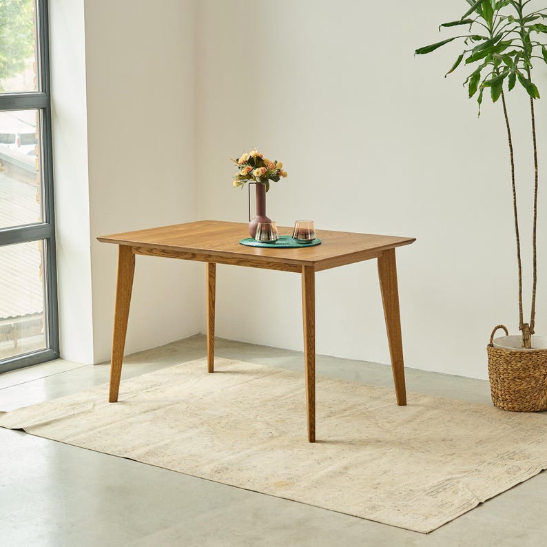 May include: A rectangular wooden dining table with four tapered legs and a smooth, light brown finish. A dark vase containing a bouquet of flowers and two small glass tumblers sit on a circular green placemat in the center of the table surface.