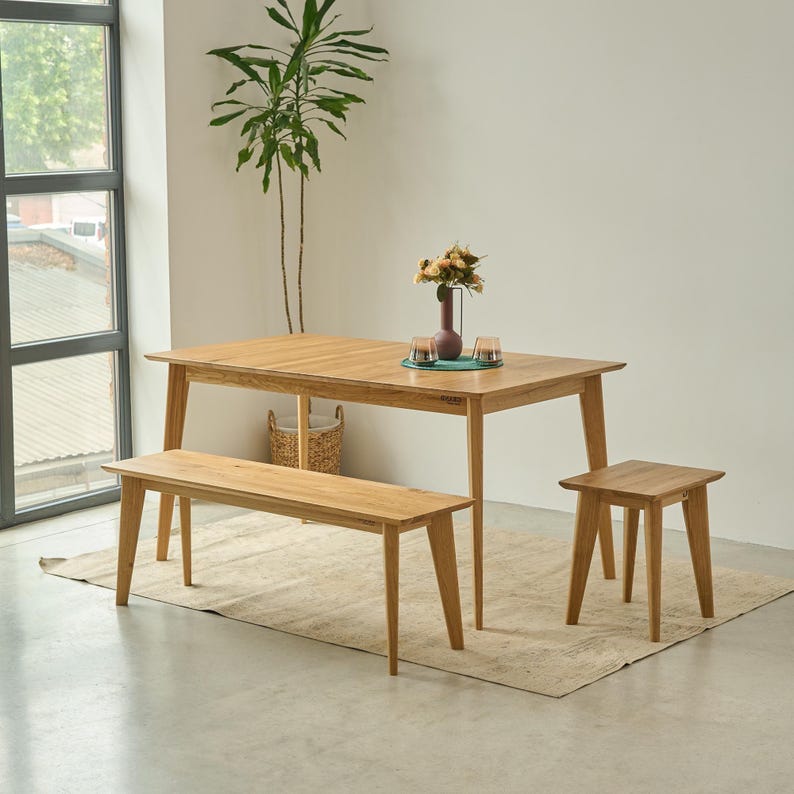 May include: A light brown wooden dining set consisting of a rectangular table with four tapered legs, a matching long bench, and a single stool. A dark vase with flowers and two small glass candle holders sit on the center of the table surface.