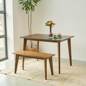 May include: A rectangular wooden dining table with four tapered legs and a matching wooden bench. The table surface holds a tall maroon vase containing a bouquet of yellow and orange flowers, alongside two small glass candle holders on a teal mat.