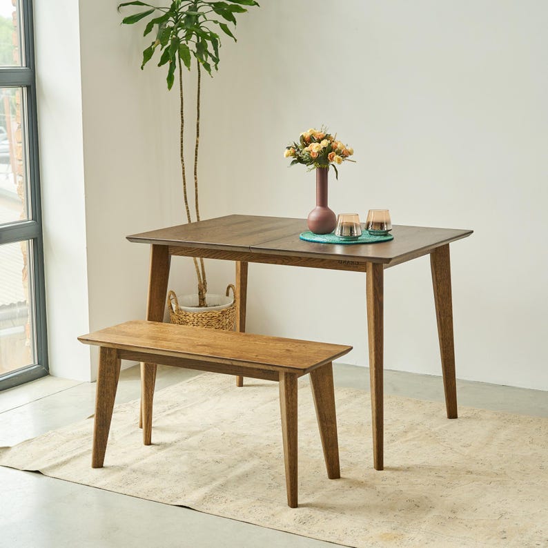May include: A rectangular wooden dining table with four tapered legs and a matching wooden bench. The table surface holds a tall maroon vase containing a bouquet of yellow and orange flowers, alongside two small glass candle holders on a teal mat.