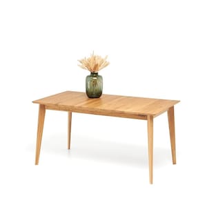 May include: A rectangular wooden dining table with four angled legs. A green glass vase with dried flowers sits on the table. The table is made of light-colored wood.