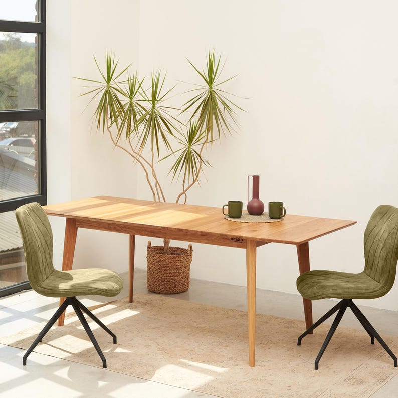 May include: A rectangular light wood dining table with four tapered legs, accompanied by two olive green upholstered chairs featuring black metal swivel bases. On the table surface sit two green mugs and a tall, slender maroon ceramic vase.