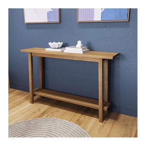 Puede incluir: Una consola de madera marrón claro con un estante inferior. La mesa es rectangular y de diseño sencillo. En la mesa se exhiben objetos decorativos, como un cuenco blanco, libros y velas. El fondo es una pared azul con obras de arte enmarcadas.