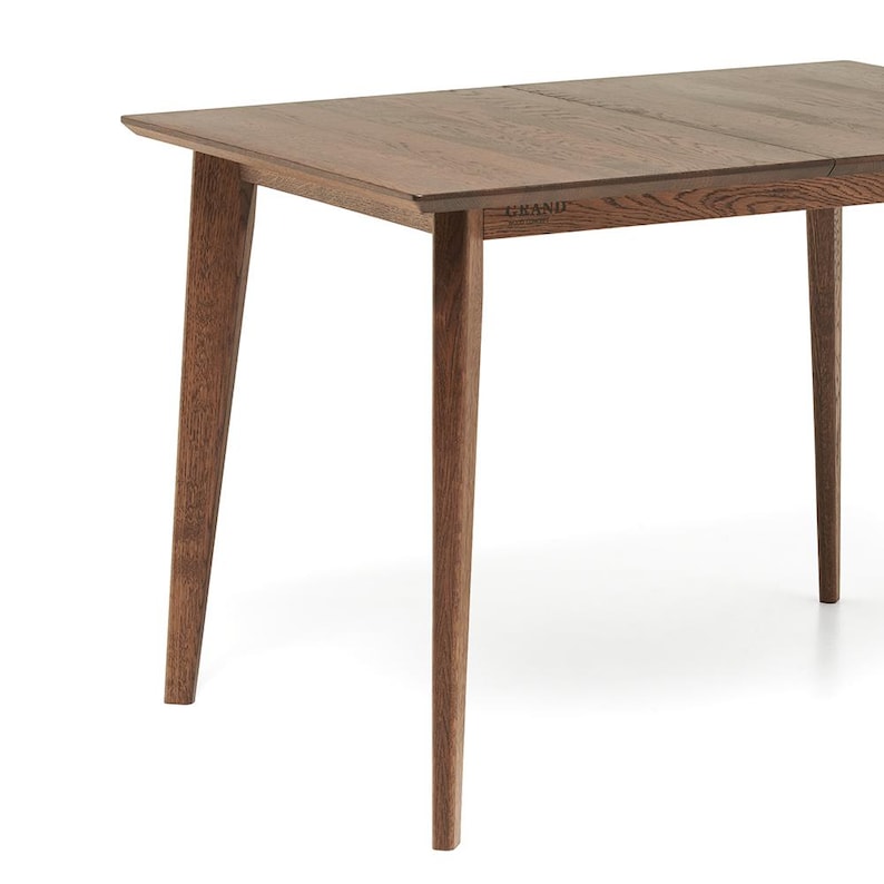 May include: A rectangular wooden table with a natural brown finish. The table has four tapered legs and a simple, modern design. The word "GRAND" is visible on the side of the table.