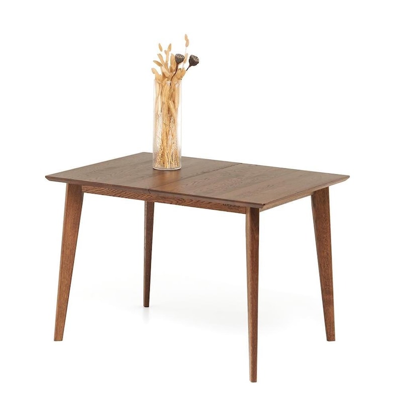 May include: A rectangular wooden dining table with four angled legs. The table is a medium brown color and has a smooth surface. A clear glass vase with dried flowers sits on top of the table.