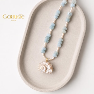 Op de afbeelding: Een ketting met een schelpenhanger en een streng lichtblauwe en witte kralen. De ketting wordt gepresenteerd op een lichtbeige ovale tray. De merknaam "Gauste Maison" staat in gouden letters linksboven.