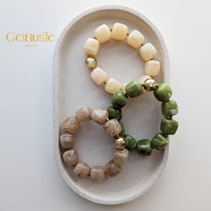 Peut inclure: Trois bracelets élastiques composés de perles lisses en forme de cube dans des tons crème, vert sauge et beige marbré. Chaque bracelet est agrémenté de petites perles métalliques dorées entre les cubes. La marque Gaëuste Maison est visible.